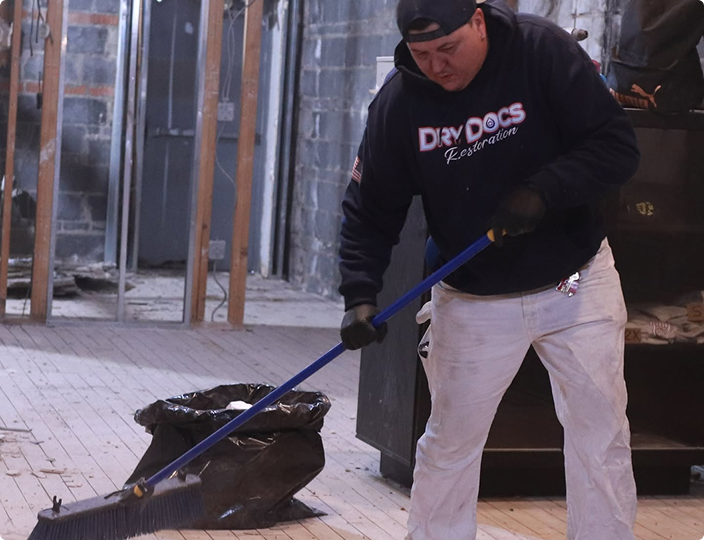 Restoration worker in a navy “Dry Docs Restoration” hoodie sweeping debris into a trash